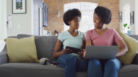 Happy-african-american-mother-and-daughter-sitting-on-sofa-using-laptop-and-tablet