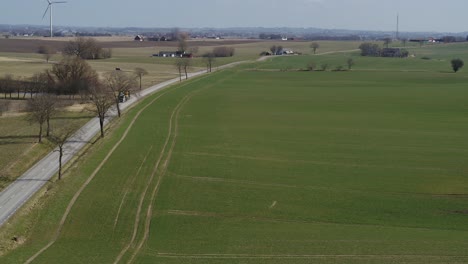 drones fly over a green field next to a road with a wind wheel in the background