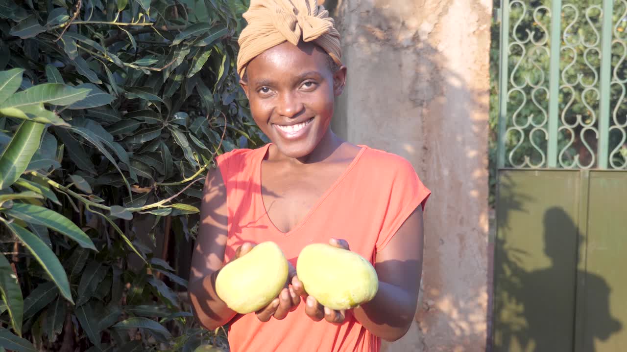 Seorang wanita Afrika mengangkat dua mangga yang baru dipetik dan tersenyum ke kamera