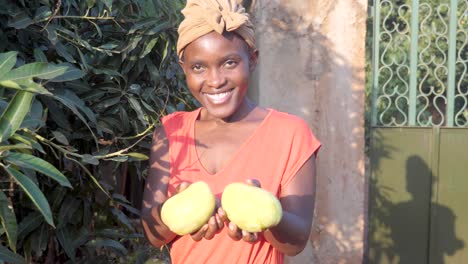 an african woman holding up two freshly picked mangoes and smiling at the camera