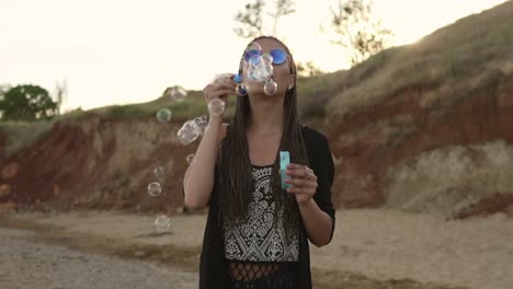 Young-female-hipster-with-dreads-cheerfully-making-soap-bubbles-on-the-beach-in-the-evening.-Slow-Motion-shot