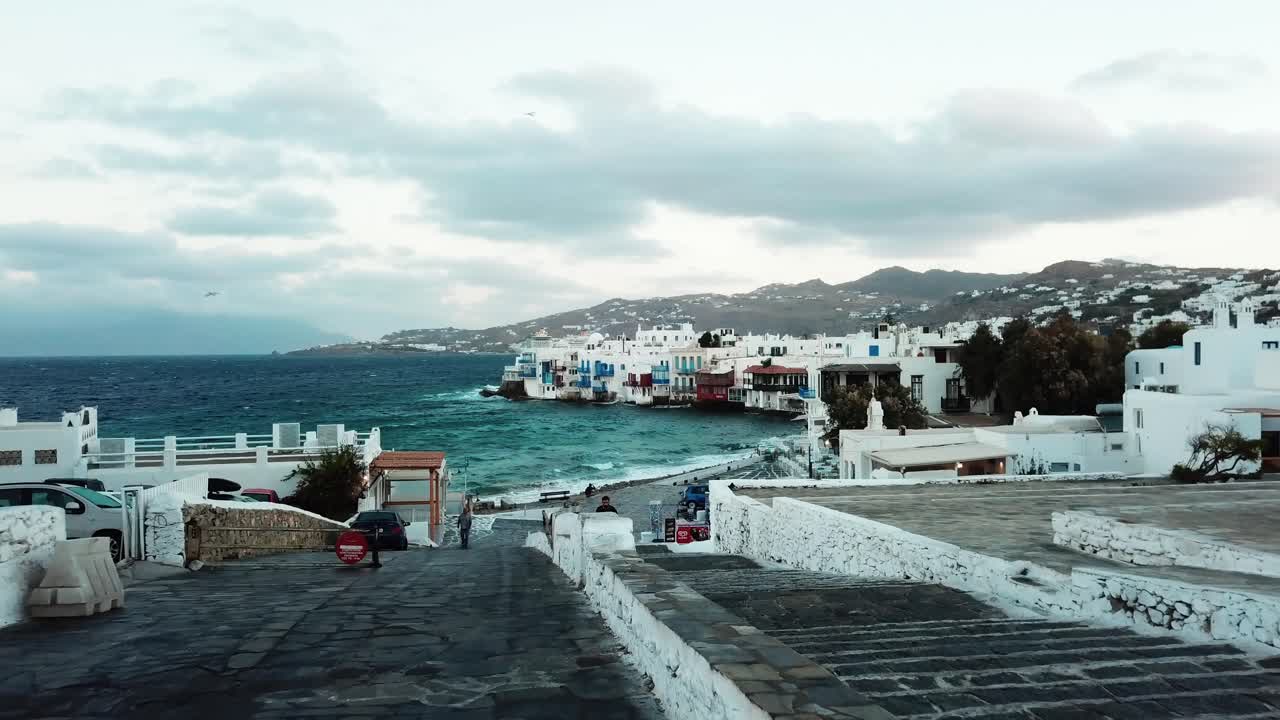 vista aérea de drones de olas de agua claras que tocan la costa con casas tradicionales en la costa con un hombre caminando en las calles de mykonos en grecia en un día nublado