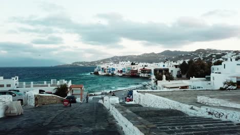 Aerial-drone-view-of-clear-water-waves-touching-the-coast-with-traditional-houses-on-the-coast-with-man-walking-in-the-streets-of-Mykonos-in-Greece-on-a-cloudy-day