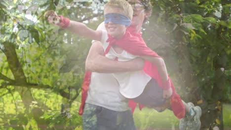 animation of foliage over happy caucasian father and son having fun dressed like superheroes