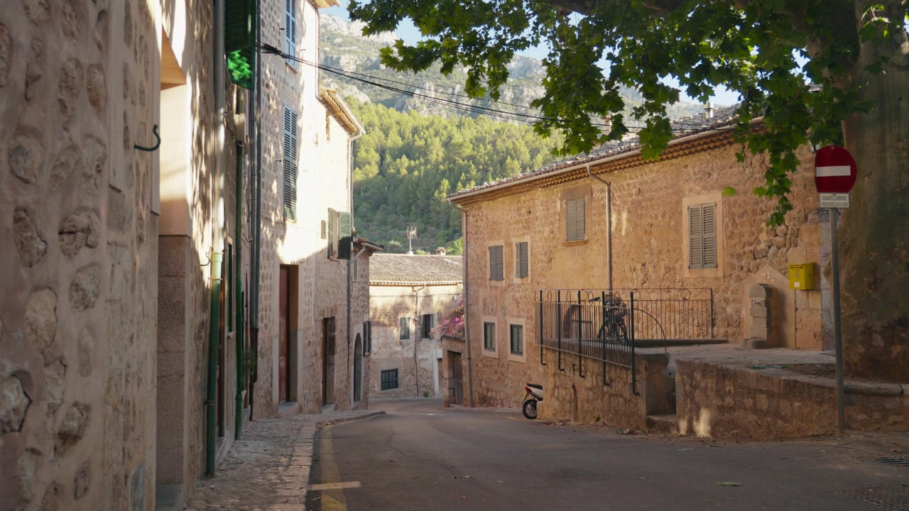 Una strada tortuosa tra case in pietra dorata, montagne boscose che si ergono sullo sfondo sotto il sole mattutino. Scena invitante di un villaggio mediterraneo per contenuti di viaggio. Campo lungo