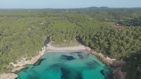 Bewaldete-Naturlandschaft,-Cala-Mitjana-Menorca-Strand-Weißer-Sand-Blaues-Meer-Drohnen-Luftaufnahme