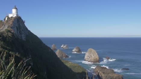 the nugget point lighthouse on top of a steep rocky highland with clumps of rocks beneath surrounded by blue ocean in catlins, new zealand - medium shot