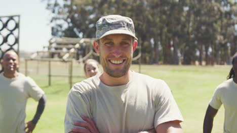 Portrait-of-caucasian-male-soldier-in-cap-smiling-at-obstacle-course-with-diverse-group-behind-him