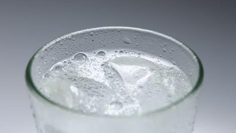 Primer-Plano-Fijo-De-La-Parte-Superior-De-Un-Vaso-Lleno-De-Refresco-Con-Gas-En-Un-Vaso-Con-Hielo,-Con-Un-Fondo-Blanco-Gris-Liso