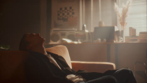 young man walking in softly lit room, leaning back on couch in deep thought