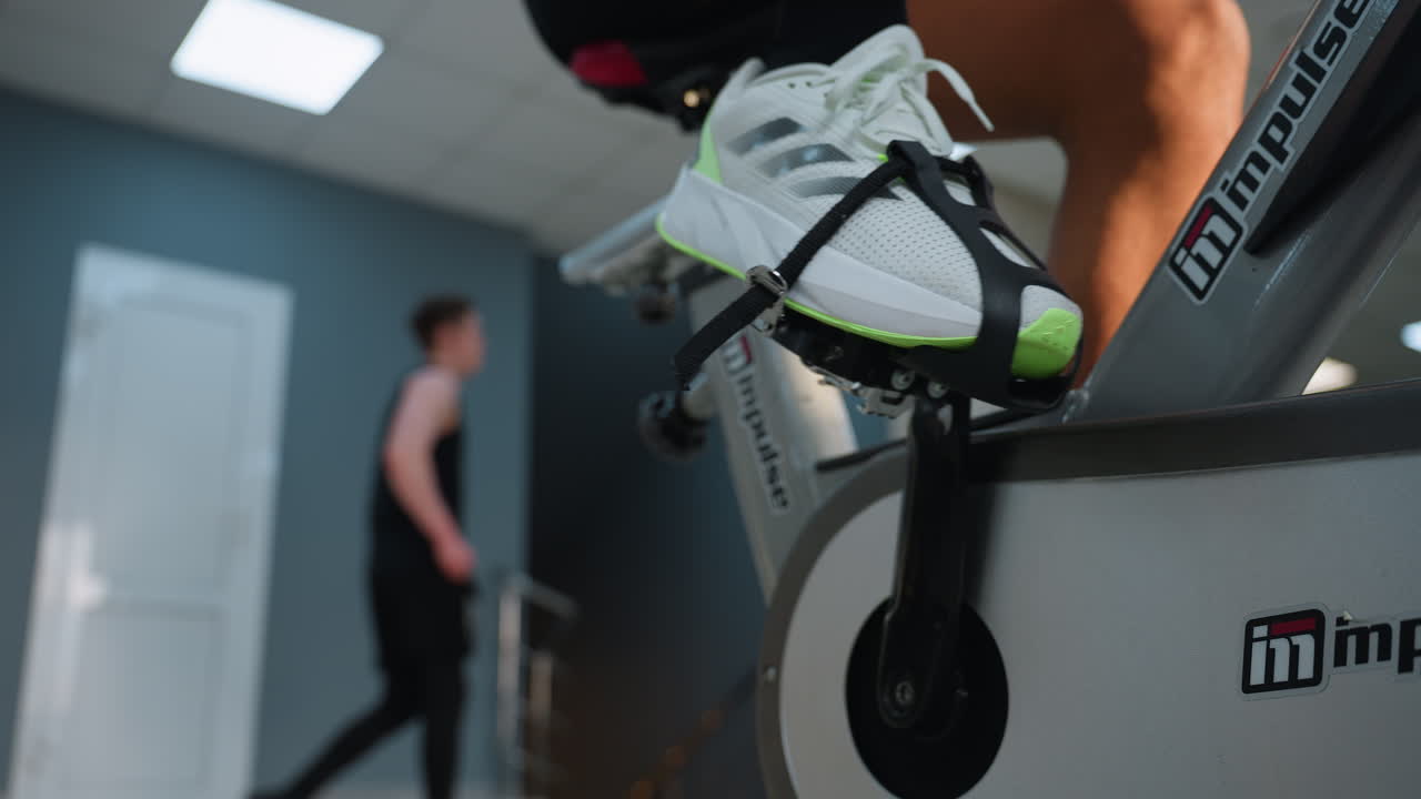 Leg view of man pedaling on impulse stationary bike in gym, showing branded frame and foot strap, with someone blurred walking behind under bright ceiling lights and modern workout environment