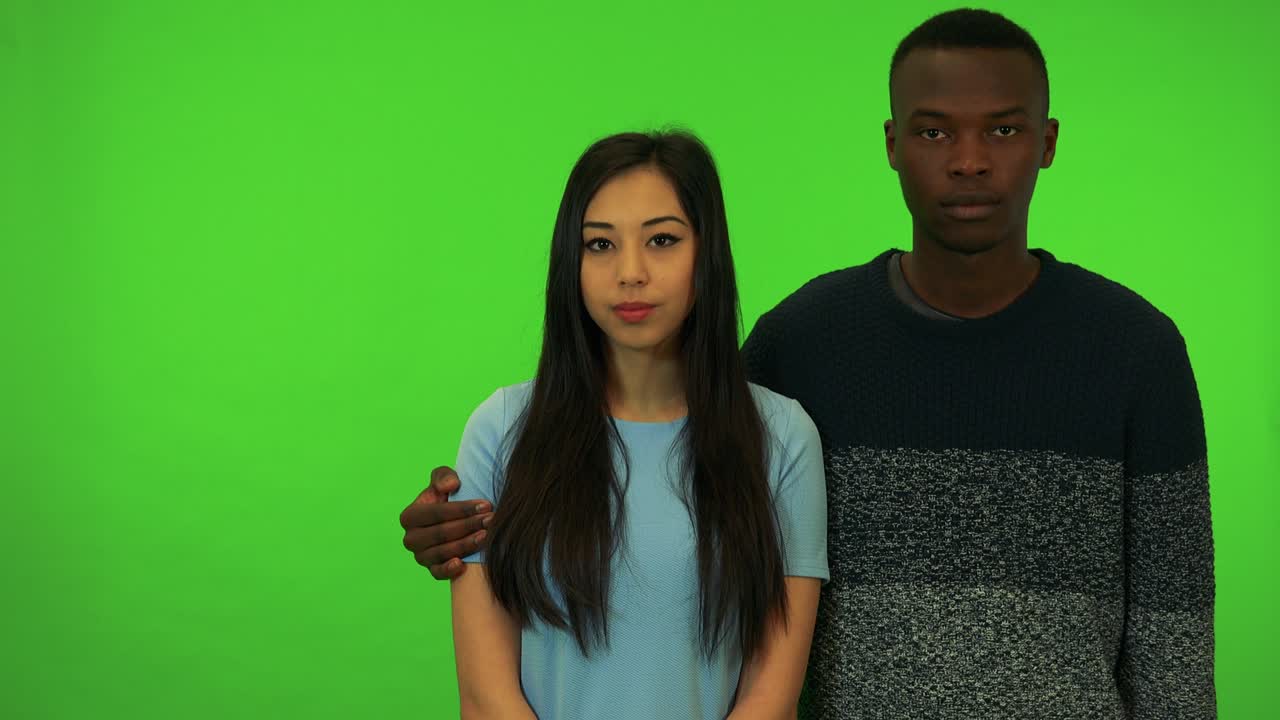 A young black man and a young Asian woman look at the camera, his arm around her shoulders - green screen studio
