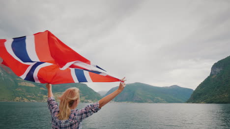 Eine-Aktive-Frau-Mit-Einer-Großen-Norwegischen-Flagge-Steht-Am-Bug-Eines-Kreuzfahrtschiffes-Fjordkreuzfahrt-In