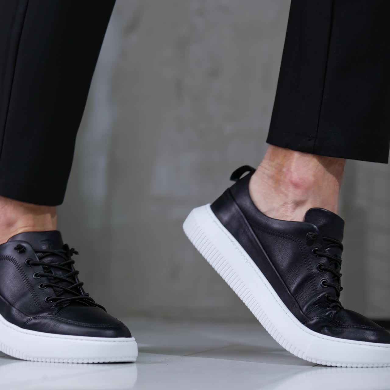 Model presenting black leather sneakers on white soles. Fashionable sport casual footwear demonstration in studio. Grey backdrop