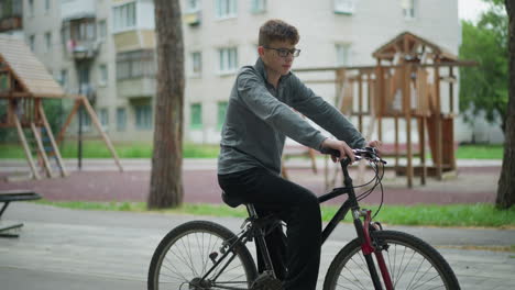 boy in a grey top and black trousers sits on his bicycle in a park setting, he is resting while holding the bike handlebars, the background features a playground structure and apartment buildings