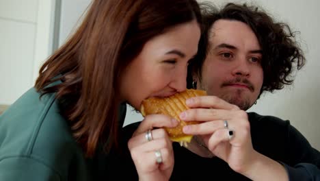 a brunette girl in a green jacket bites a sandwich that her boyfriend is holding in his hands who wanted to eat the sandwich himself at home