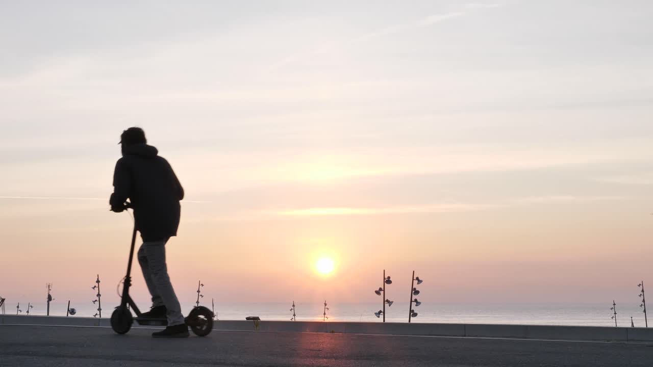 Joven moderno conduciendo patinete eléctrico se detiene para ver el amanecer en paseo marítimo de hormigón