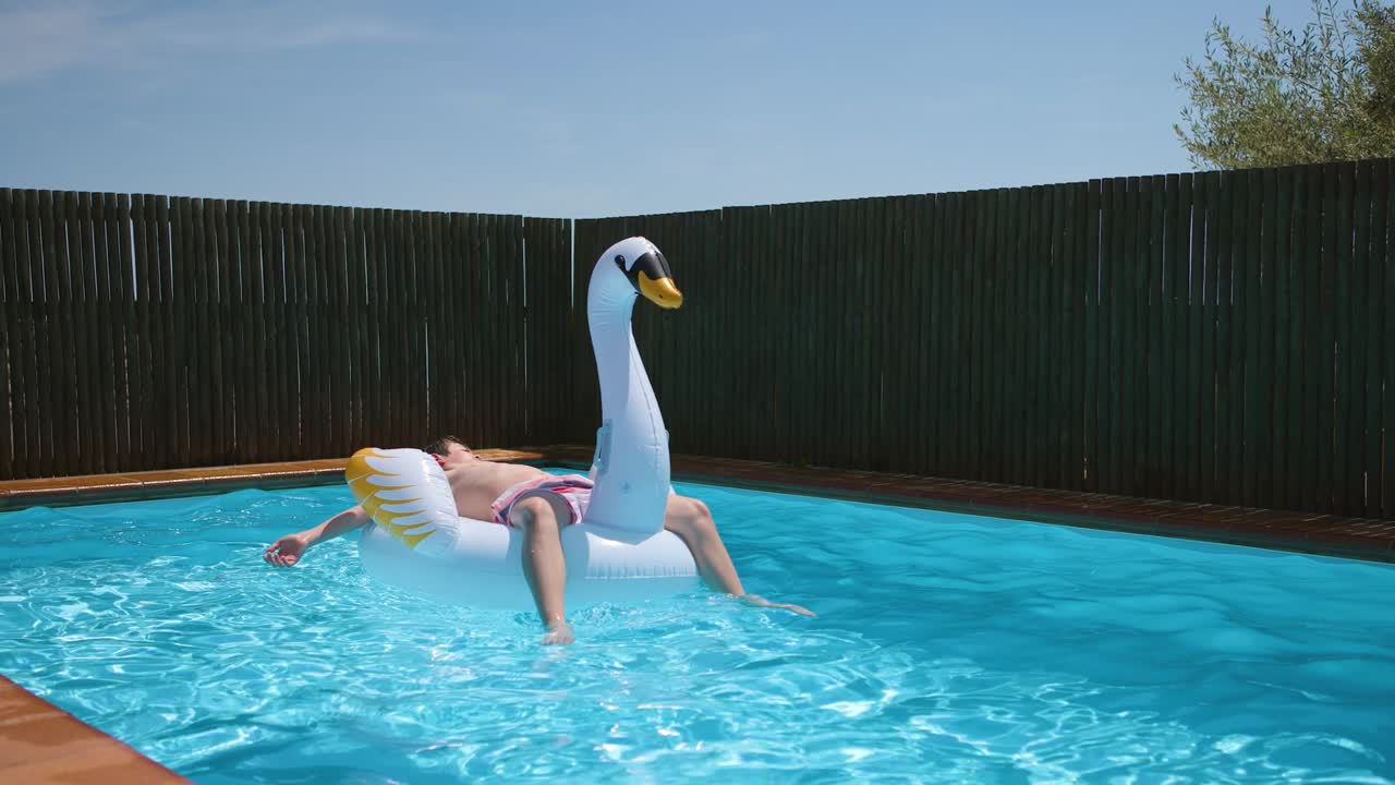 Teen boy relaxes and sunbathe on inflatable swan toy in blue pool