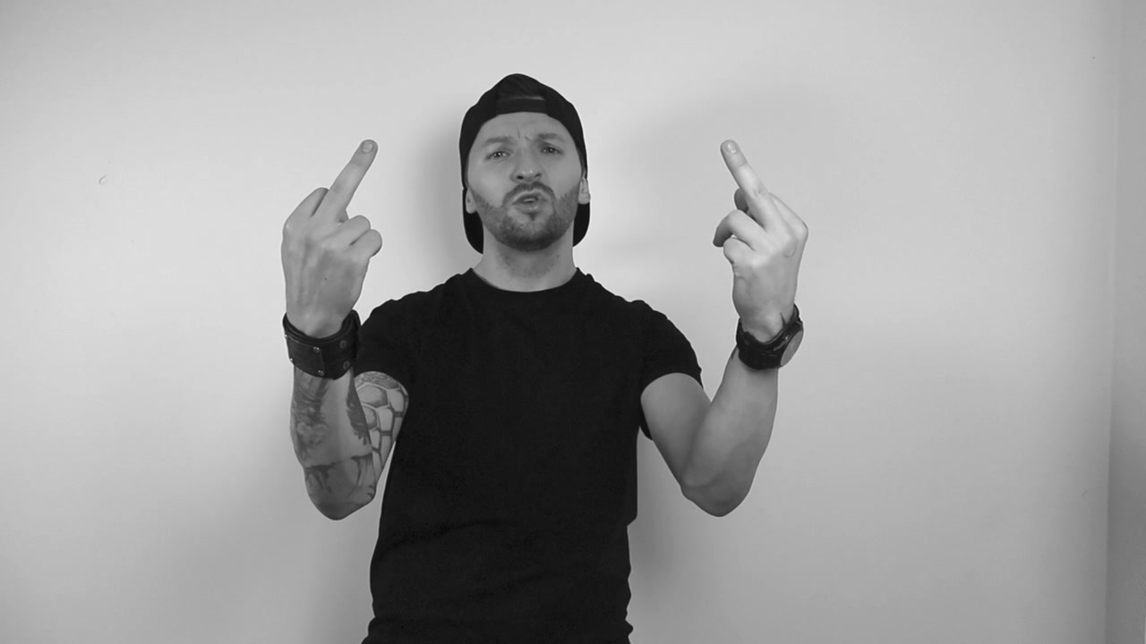Grayscale footage of a tattooed man flipping a person off by showing middle finger directly at the camera in slow motion. Man is lit by studio lights and is standing. Anger and bad mood insulting.