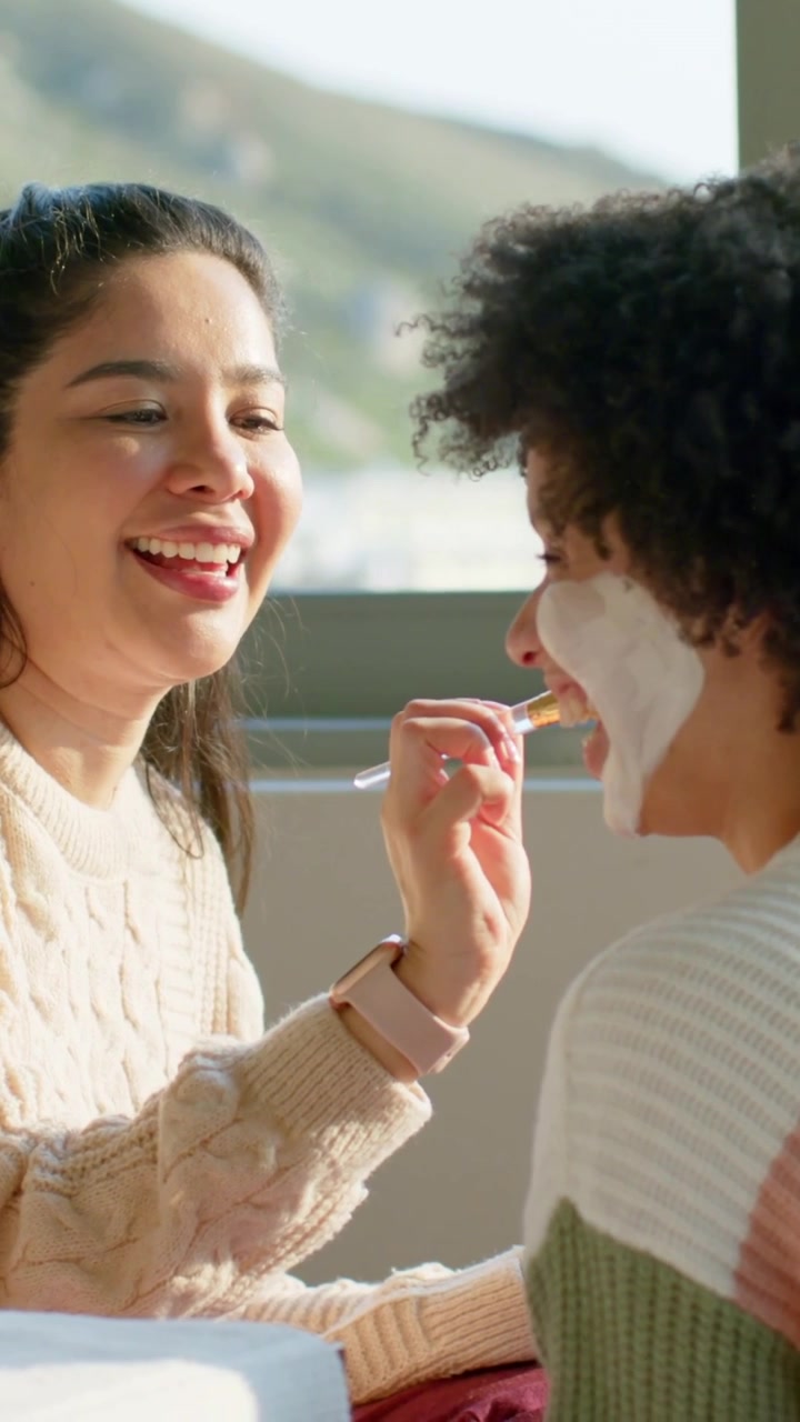 Video verticale: Sorridendo, una donna con top color crema applica una maschera all'amica, alla finestra con un barattolo per la cura.