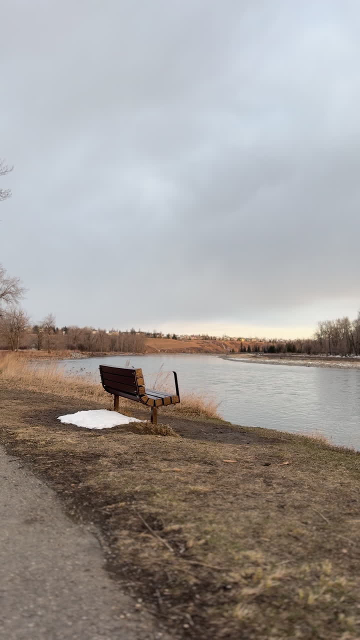 Una panchina di legno del parco si affaccia su un ampio fiume delimitato da erba secca e alberi in lontananza. La luce del primo tramonto e il cielo aperto creano un'ambientazione fluviale tranquilla e contemplativa