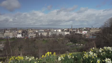 Panning-shot-of-Princes-Street-Gardens-and-city-of-Edinburgh-with-white-and-yellow-daffodils-in-the-foreground-on-a-nice-sunny-day-with-nice-clouds-in-Scotland