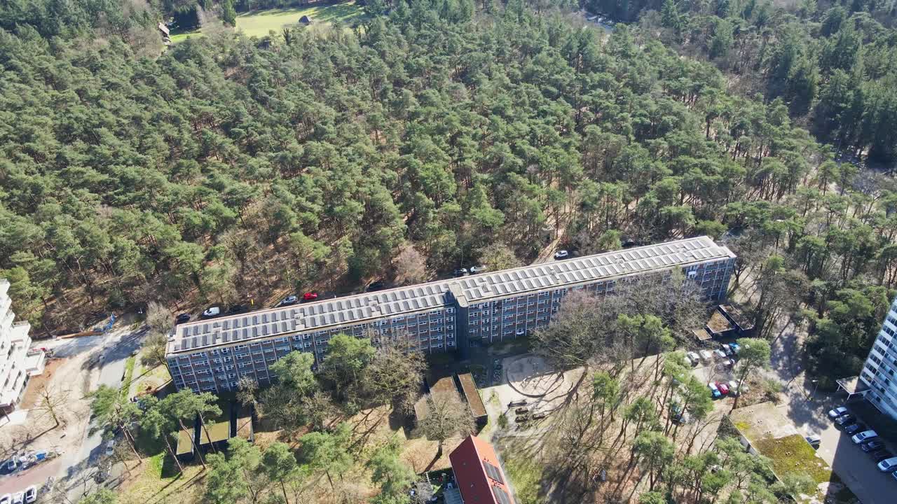 Beautiful aerial tilt down from a stunning green forest to a modernized apartment building with a rooftop filled with solar panels on a sunny day