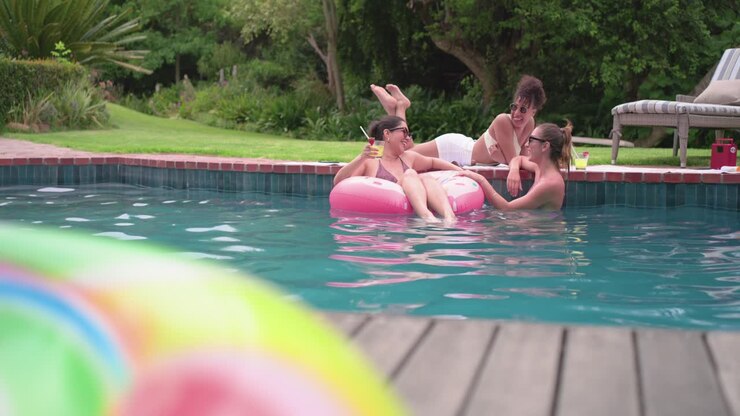 Diverse women in bikinis reaching and passing yellow cups on pink float in backyard pool, toasting