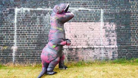 Divertido-Disfraz-De-Dinosaurio-Prehistórico-Deteniéndose-Y-Mirando-Hacia-Arriba-Junto-A-La-Pared-De-Ladrillo