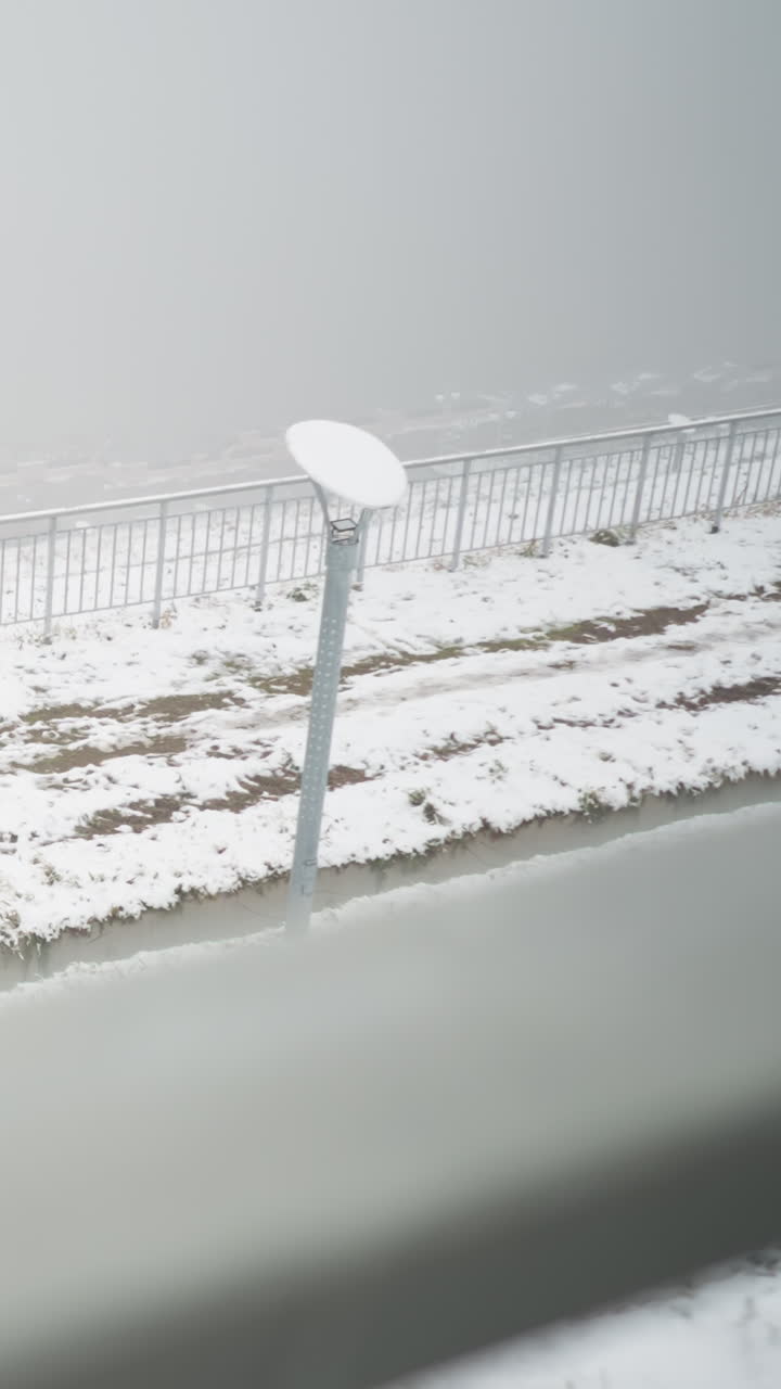 Snow-covered landscape cloaked in dense fog, metal railings and light pole above brown soil and patchy snow, embankment slope and path receding into mist