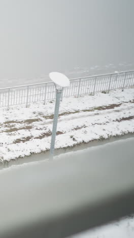 snow-covered landscape cloaked in dense fog, metal railings and light pole above brown soil and patchy snow, embankment slope and path receding into mist