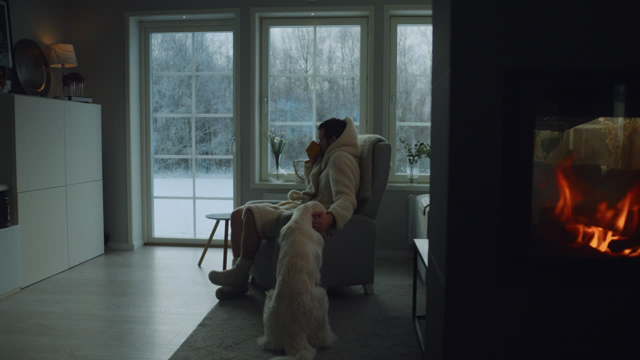 Man with his dog relaxing in winter at home with a cup of tea and fireplace.