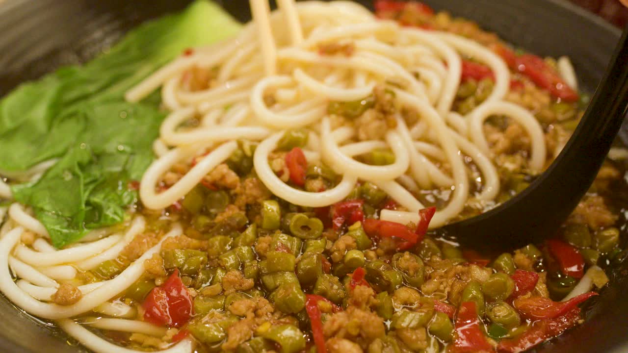 Chopsticks lift thick noodles from spicy broth with bok choy, minced meat, and chili peppers