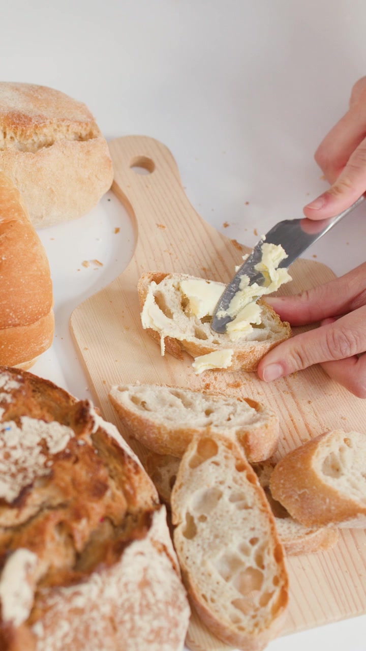 Hands spread creamy butter on baguette slices atop a wooden board with various artisan breads.