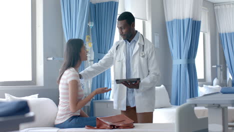 diverse male doctor using tablet and female patient, talking in hospital ward, slow motion