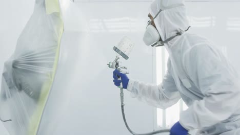 mixed race male car mechanic with protective clothes and using a spraying gun on a piece of a car