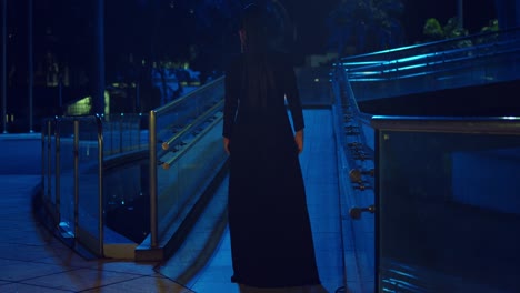 hispanic woman walks on a walkway in the city in a black dress and blue lights in the background