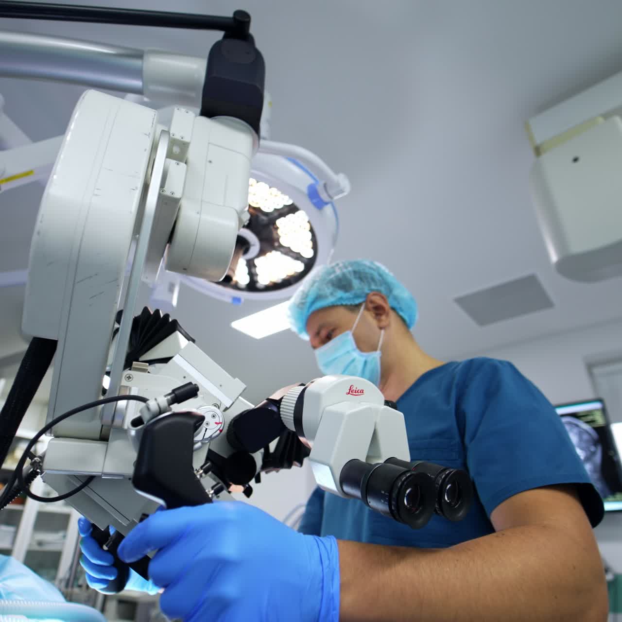 Un médico uniformado traslada el equipo en un quirófano moderno. Un doctor ajusta el potente microscopio sobre el paciente en la mesa de operaciones. Vista en ángulo bajo.