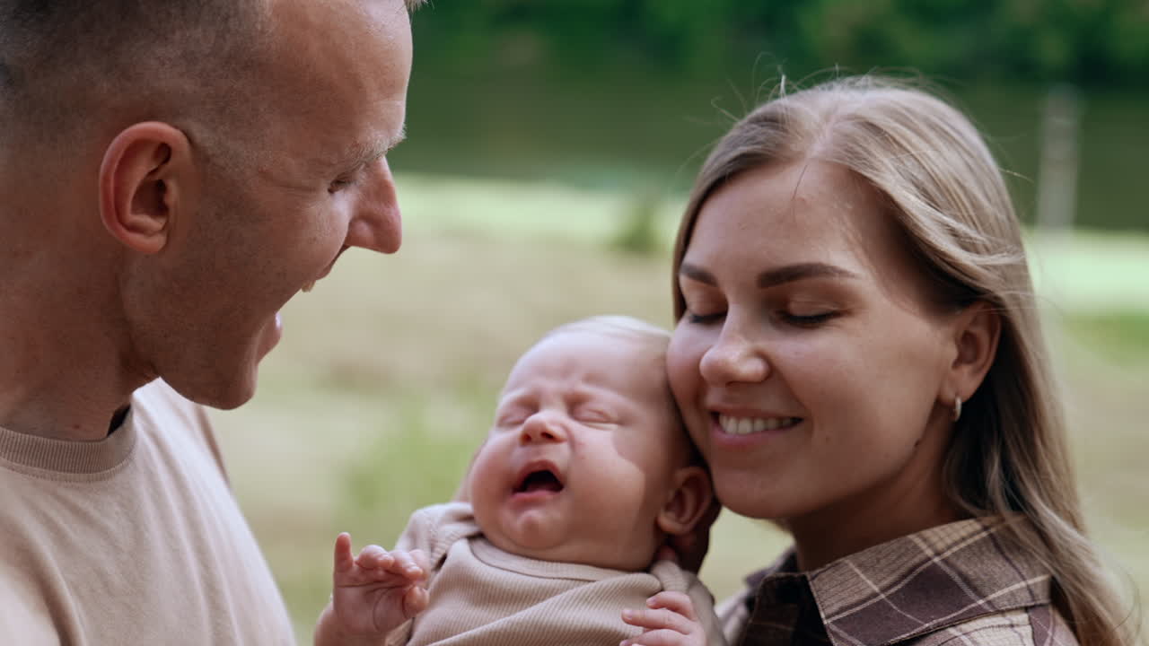 Loving Caucasian parents kiss their newborn from two sides. Little baby yawns and waves tiny hands. Blurred nature backdrop.