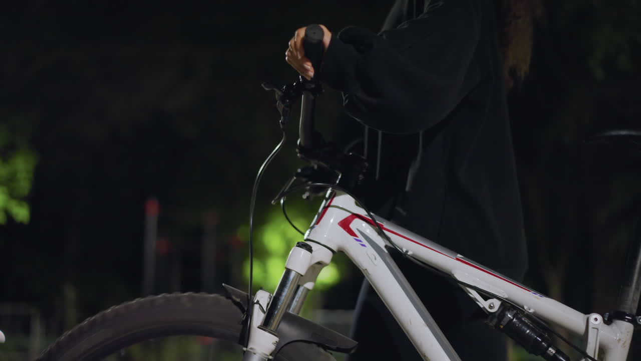 Park Bike Headlight Check Under Lamp, Person In Dark Hoodie Adjusts Front Light And Brake Cables, CloseUp Of Handlebars And Frame With Soft Green Background, Careful Preparation For Night Ride
