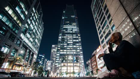 a man sits on the sidewalk in a bustling urban area at night. he appears contemplative, surrounded by towering skyscrapers and bright city lights.