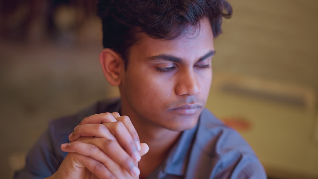 Young man sitting with clasped hands, looking slightly down and away in quiet indoor setting, showing calm expression, introspective emotion, and still posture, capturing thoughtful mood and reflective energy