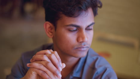 young man sitting with clasped hands, looking slightly down and away in quiet indoor setting, showing calm expression, introspective emotion, and still posture, capturing thoughtful mood and reflective energy