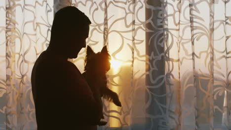 A-Young-Single-Man-Standing-At-The-Window-Holding-His-Dog-Pensively-Looking-Into-The-Window-At-Sunse