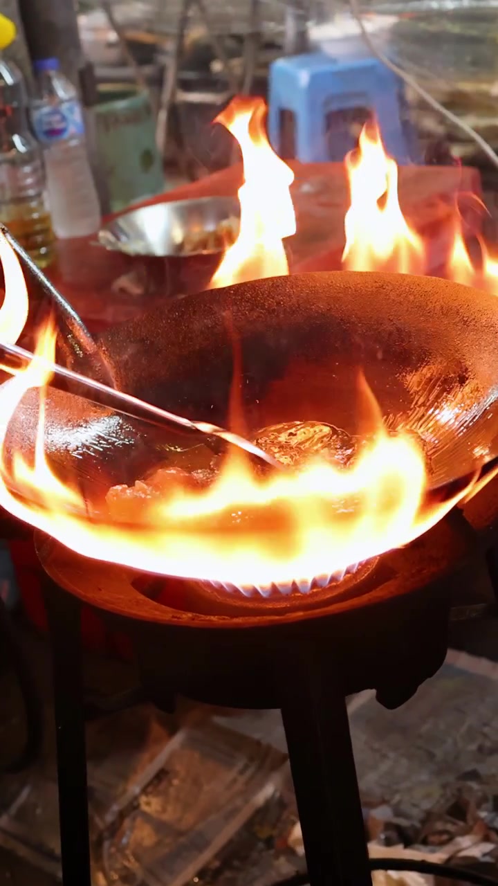 Wajan di atas api terbuka yang intens, masakan makanan jalanan, suasana Bangkok yang semarak, kamera genggam, pencahayaan alami