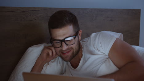 vista de cerca de un hombre sonriente usando una laptop y sonriendo mientras ve algo por la noche en su cama