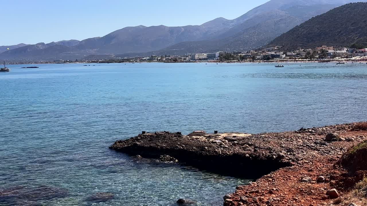 Calm sea with a rocky shore, distant beach, and mountains in Crete Greece under a clear sky
