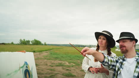 painter wearing a checkered shirt and hat is pointing with a brush,to a woman beside him, dressed in a white dress and hat. they are in a grassy field with a partially painted canvas in the background