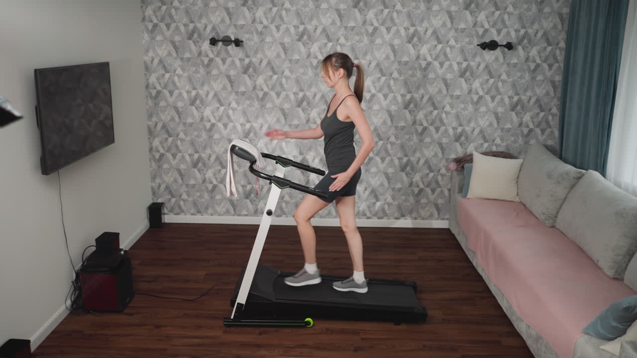 Woman walking on treadmill indoors, swinging one arm naturally while stepping forward, dressed in dark workout outfit, towel hanging on handlebars, cozy living room with sofa and curtain