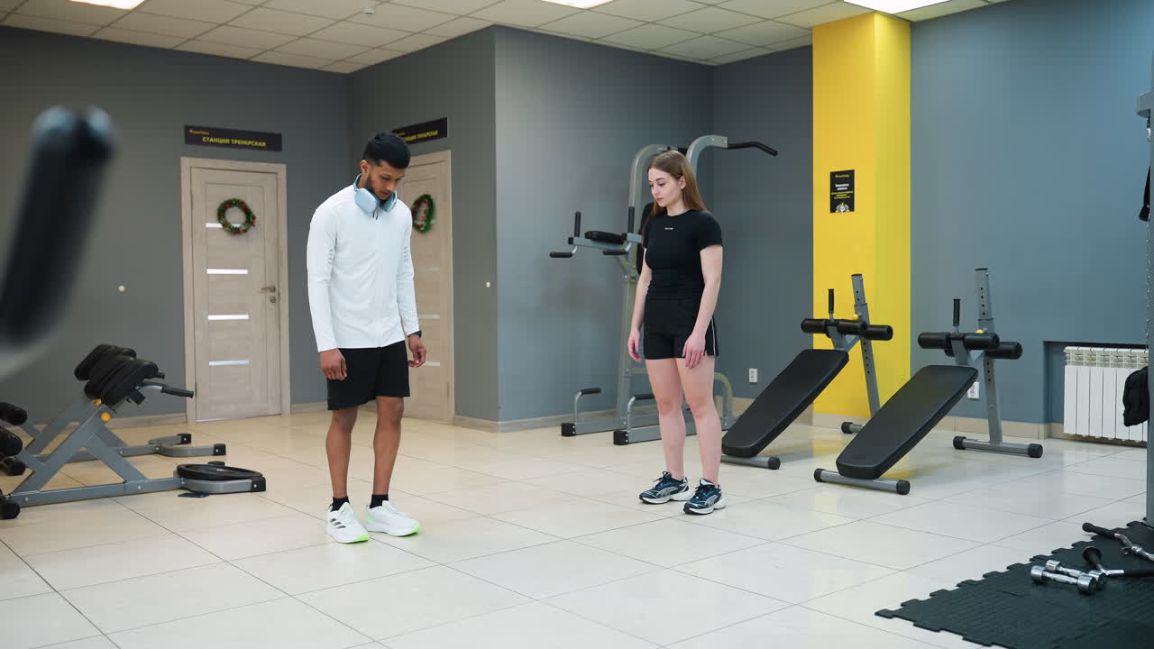 gym instructor in white jacket stands in fitness room demonstrating kneel squat technique to young woman in sportswear with modern workout equipment and gym furnishings visible in background